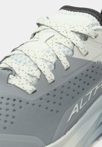 Gros plan sur une chaussure de sport gris clair et blanche avec des lacets à motifs et le nom de la marque "Altra" sur le côté.