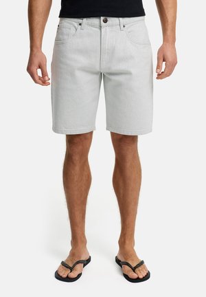 Mann kledd i lysegrå denimshorts og svarte flip-flops, stående mot en ensfarget hvit bakgrunn.