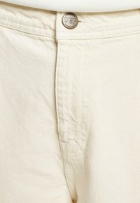 Helle beige Jeans mit einem Metallknopf, auf dem "DEF" eingraviert ist, robusten Nähten und glatter Textur.