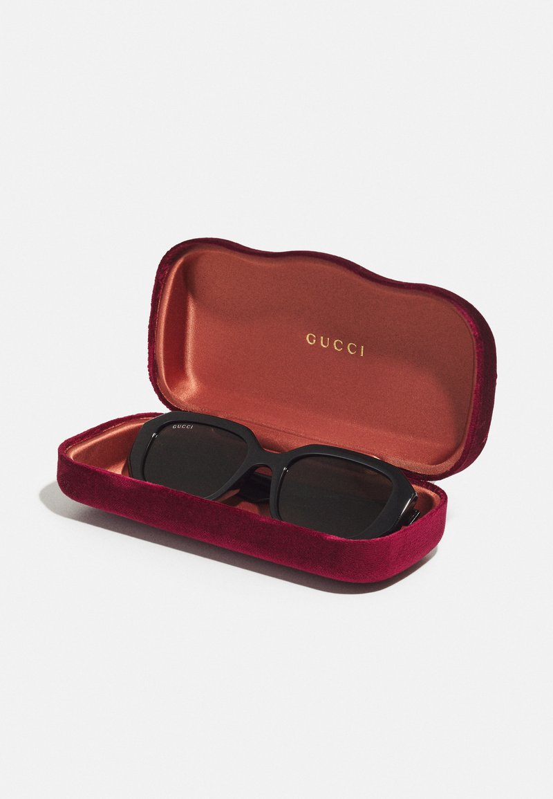 Gucci-Sonnenbrille in einem schwarzen Etui, innen in einer bordeauxroten Samtbox mit goldenem "GUCCI"-Logo auf einem orangefarbenen Innenfutter.
