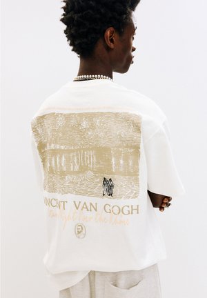 Witte t-shirt met een Van Gogh-print in beige, dat een scène met twee figuren afbeeldt. De tekst eronder luidt "Vincent Van Gogh" in goud.