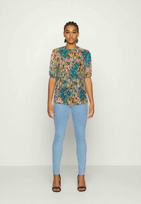 Top de manga curta, multicolorido, com padrão florido em tecido leve; combinado com jeans skinny azul claro e sandálias de salto alto pretas.