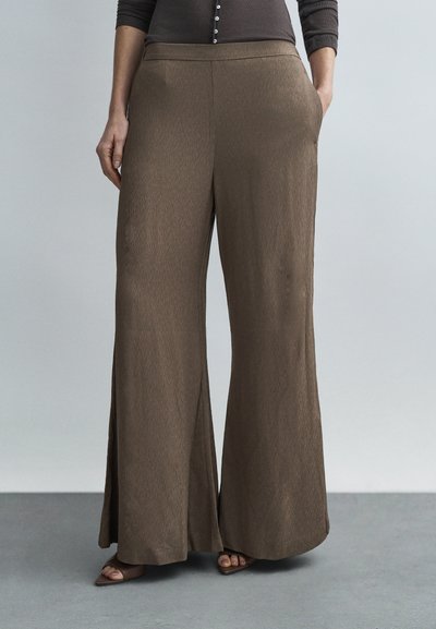Femme portant un pantalon marron à taille haute et jambes larges avec poches latérales, associé à un haut foncé et des sandales marron à bouts ouverts sur un sol gris.