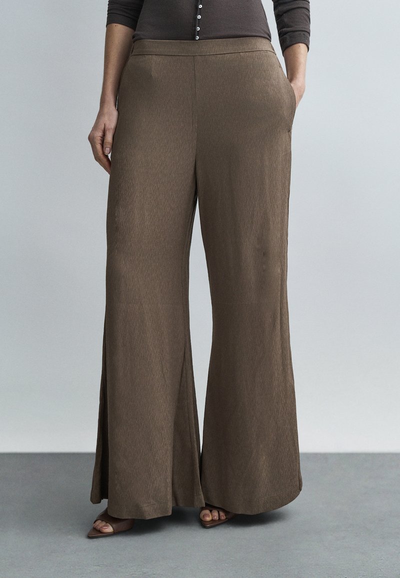 Donna che indossa pantaloni marroni a gamba larga con vita alta e tasche laterali, abbinati a una maglia scura e sandali marroni aperti sulle dita, su pavimento grigio.