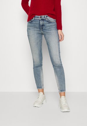 Jeans ajustados de mezclilla azul claro con detalles de desgaste, combinados con un suéter rojo texturizado y zapatillas gris claro.