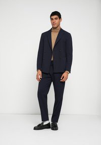 Marinblå dubbelknäppt kavaj med notched slag, bärs över en beige polotröja, kombinerad med skräddarsydda byxor och svarta skor.