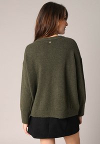 Pull en tricot vert avec une coupe ample, un col rond et des épaules tombantes. Présente un petit logo dans le dos et des bords côtelés.