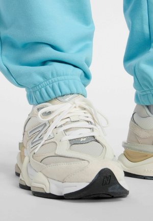 Persoon die beige en witte New Balance sneakers met witte veters en lichtblauwe joggingbroek draagt, staande op een witte ondergrond.