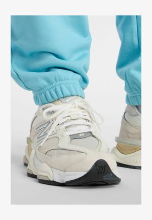 Persoon die beige en witte New Balance sneakers met witte veters en lichtblauwe joggingbroek draagt, staande op een witte ondergrond.
