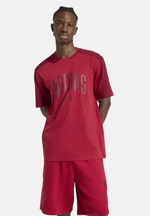 Jeune homme debout, les mains dans le dos, portant un t-shirt et un short rouges assortis Adidas sur un fond clair uni.