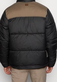 Giubbotto imbottito nero con una parte superiore marrone, caratterizzato da cuciture orizzontali, una siluetta cropped e un'etichetta con logo sul retro.