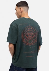 T-shirt oversize vert foncé en coton, avec un grand graphisme d'œil rouge et des accents floraux au dos, doté d'un col rond simple.