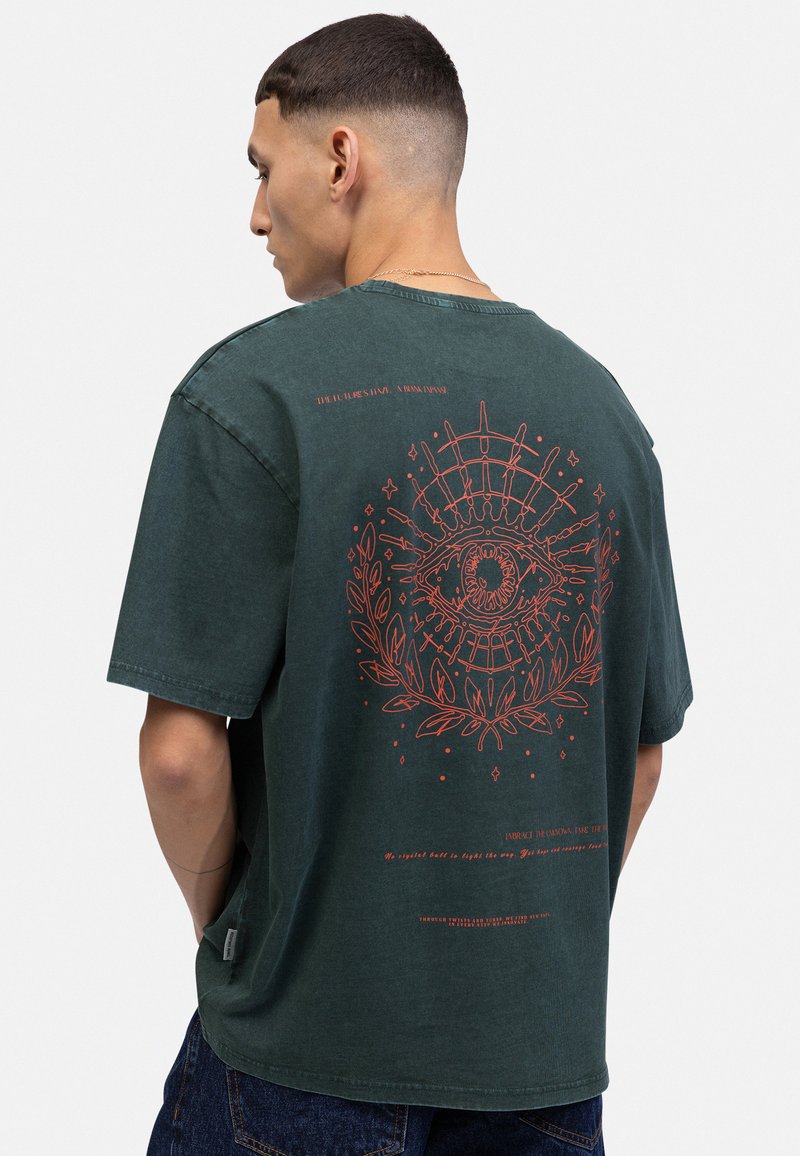 T-shirt oversize vert foncé en coton, avec un grand graphisme d'œil rouge et des accents floraux au dos, doté d'un col rond simple.