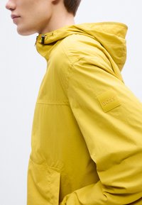 Κίτρινο αδιάβροχο μπουφάν με κουκούλα, που διαθέτει μπροστινή τσέπη και τετράγωνο λογότυπο "BOSS" στο μανίκι. Κατασκευασμένο από λείο, ελαφρύ υλικό.