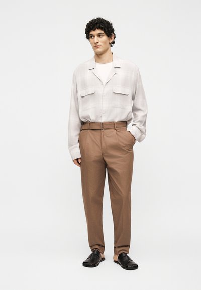 Άνδρας με σγουρά μαλλιά που φοράει ανοιχτό μπεζ πουκάμισο με κουμπιά, καφέ ψηλόμεσο πλισέ παντελόνι και μαύρα πλεκτά σανδάλια, στέκεται μπροστά σε απλό φόντο.