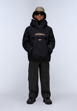 Schwarze Nylon-Pulloverjacke mit einem Logo-Patch und norwegischer Flagge. Kombiniert mit olivgrünen Cargo-Hosen und schwarzen gemusterten Schuhen, accessoriert mit Sonnenbrille und einem beigen Bucket Hat.