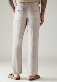 Pantaloni di lino beige chiaro con una vestibilità dritta, due tasche posteriori con bottoni scuri e texture ruvida. Indossati con scarpe basse.