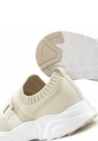 Beige Sportschuhe mit einem gestrickten Obermaterial, einer strukturierten Mittelsohle und einer gemusterten Gummisohle. Einfaches Design mit einer Schlaufe an der Ferse.