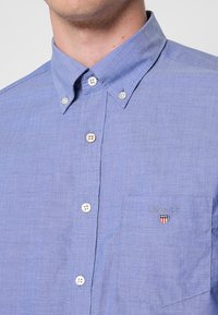 Hellblaues Hemd mit Knopfleiste aus strukturiertem Stoff, ausgestattet mit einer Tasche mit Logo und weißen Knöpfen an einem klassischen Kragen.
