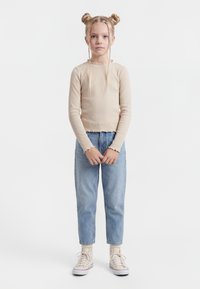 Jauna meitene ar gaišiem matiem sasietiem bulciņās un bizēs, tērpusies bēšā garroku krekliņā, gaišzilās džinsās un baltās augstpapēžu kedās, stāv nekustīgi.