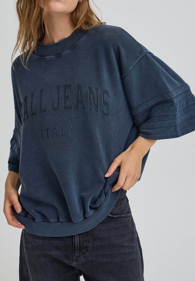 Femme portant un sweat-shirt bleu marine oversize avec le texte "ALL JEANS ITALY" et des manches mi-longues plissées, associée à un jean noir.