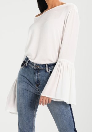 Woman wearing off-shoulder white blouse with flared sleeves and high-waisted blue jeans with dark side stripes.