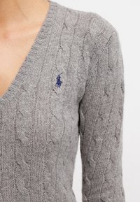 Harmaa palmikkoneule, jossa on V-kaula-aukko ja pitkät hihat. Vasemmassa rinnassa sininen brodeerattu logo. Pehmeä rakenne, keskivahvuinen.