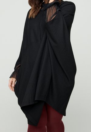 Vrouw draagt een loszittende zwarte poncho over doorschijnende zwarte mouwboorden en bordeauxkleurige legging tegen een eenvoudige achtergrond.