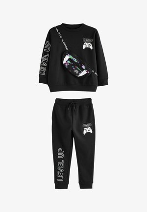 Schwarzer Trainingsanzug mit einem Langarmshirt, das weißen Text und grafische Designs aufweist, sowie passenden Hosen mit einem Kordelzug. Inklusive einer gemusterten Bauchtasche.