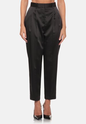 Donna che indossa pantaloni in raso neri a vita alta e gamba affusolata abbinati a décolleté nere con punta affilata, in piedi su sfondo bianco.