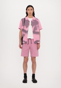Korte mouwen roze shirt met grijs abstract patroon, witte T-shirt met opdruk, roze gebloemde shorts en zwarte sokken met sandalen.