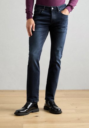 Person iført mørke, tætsiddende jeans, sorte læder snørestøvler og en lilla langærmet top, der står på et trægulv.