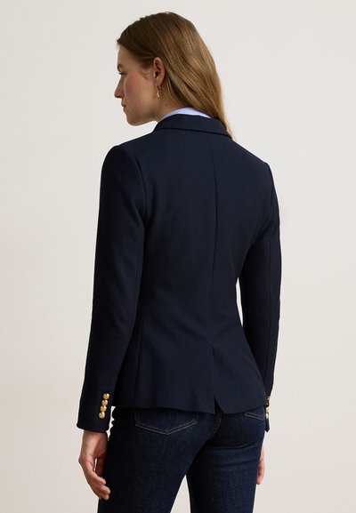 Femme vue de dos portant un blazer bleu marine sur mesure avec des boutons dorés sur les manches et un jean bleu foncé sur un fond clair uni.