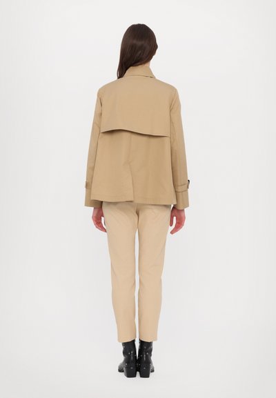 Veste marron avec un dos superposé, associée à un pantalon beige et des bottines noires. Le matériau semble lisse et structuré.