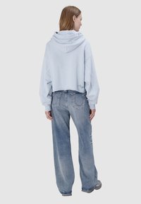 Kobieta w jasnoniebieskiej krótkej bluzie z kapturem i luźnych niebieskich dżinsach stoi tyłem na jednolitym tle.