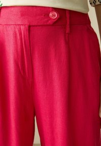 Pantalon fuchsia en tissu doux, avec un plissé à l'avant et un détail de bouton rose à la taille. La texture semble lisse.