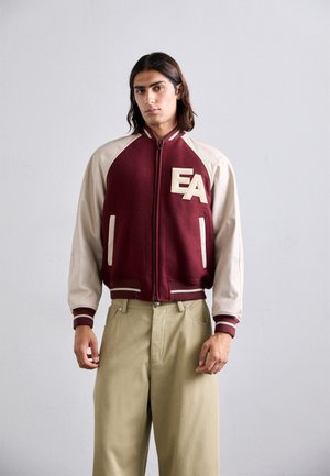 Junger Mann mit langen dunklen Haaren, der eine weinroten und beigen Collegejacke mit "EA"-Buchstaben und beigen weiten Hosen trägt.