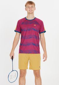 Herremode: maroon kortærmet skjorte med blåt bølgemønster, gyldne shorts. Holder en badmintonketsjer med blå detaljer.