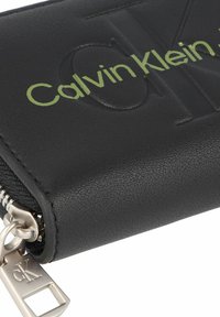 Portafoglio in pelle nera con la scritta "Calvin Klein" in verde in rilievo. Presenta una chiusura con cerniera e un dettaglio in metallo argentato con una targhetta del logo.