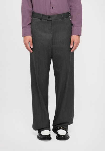 Homme portant un pantalon large gris foncé, des chaussettes blanches, des mocassins noirs avec un détail en chaîne dorée, et une chemise mauve à boutons, se tenant contre un fond uni.