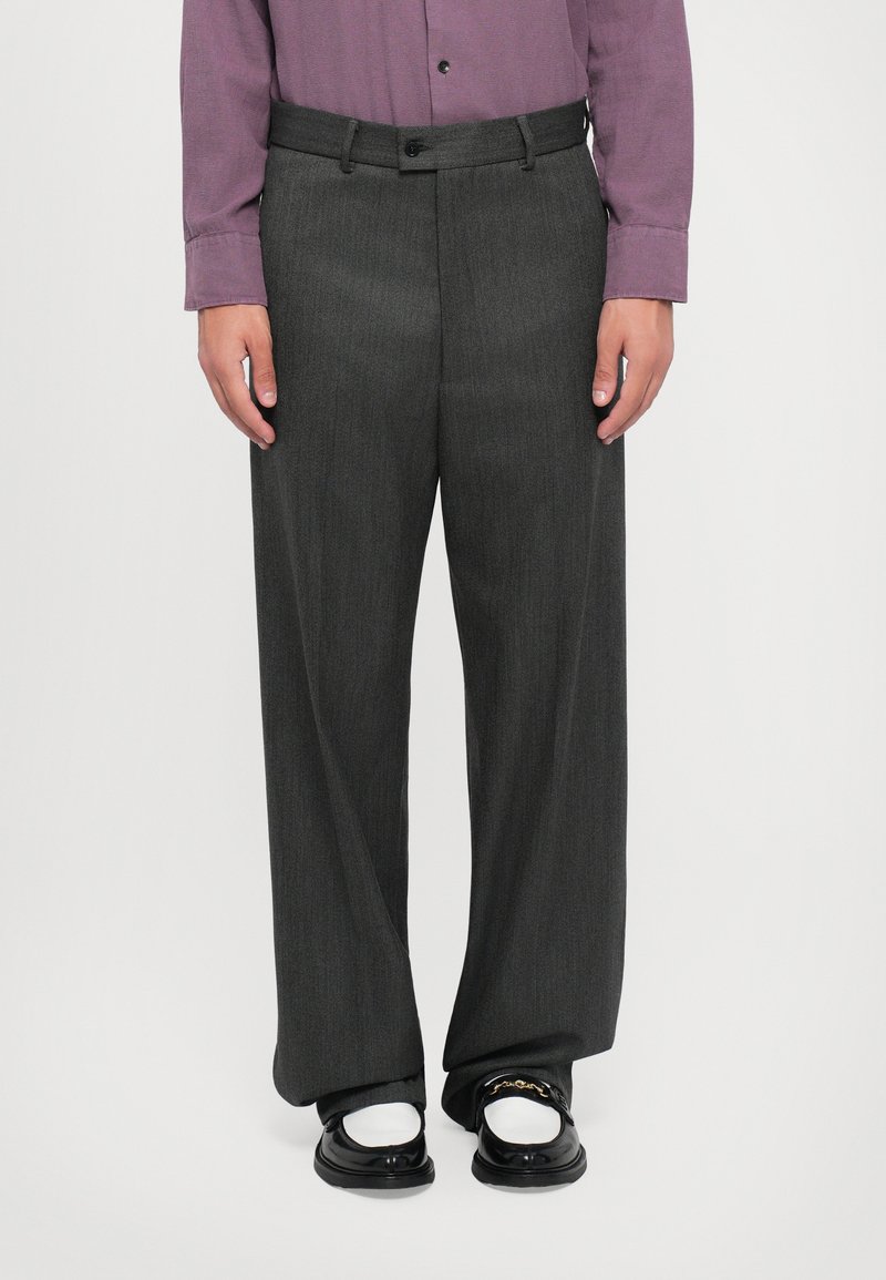 Homme portant un pantalon large gris foncé, des chaussettes blanches, des mocassins noirs avec un détail en chaîne dorée, et une chemise mauve à boutons, se tenant contre un fond uni.