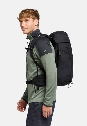 Sac à dos noir d'extérieur avec plusieurs compartiments, porté par une personne dans une veste légère verte, doté de bretelles réglables et d'une surface texturée.