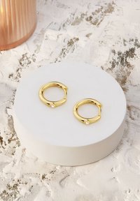 Orecchini a cerchio in oro con un design elegante e un piccolo accento a perla. Posizionati su una superficie rotonda bianca. Sfondo testurizzato.
