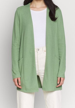 Cardigan aperto verde chiaro, realizzato in tessuto testurizzato. Presenta maniche lunghe e due tasche frontali. Indossato sopra una maglietta bianca e pantaloni chiari.
