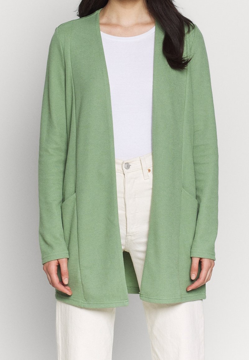 Cardigan aperto verde chiaro, realizzato in tessuto testurizzato. Presenta maniche lunghe e due tasche frontali. Indossato sopra una maglietta bianca e pantaloni chiari.