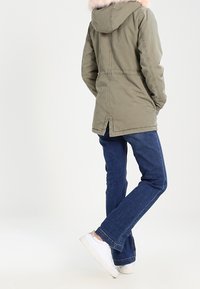 Osoba oblečená v olivově zelené parce s kapucí lemovanou růžovým kožíškem, v modrých džínách a bílých teniskách, stojí s zády otočenými vpřed.