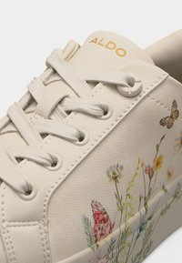 Zapatilla Aldo color crema con cordones blancos e ilustraciones detalladas de flores y mariposas en su superficie de lona.