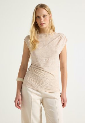 Lyshåret kvinde iført en bleg beige tekstureret kortærmet top og off-white bukser med vide ben, poserer med et neutralt udtryk.