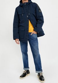 Námořnicky modrá zateplená parka bunda se zipovým uzávěrem, několika předními kapsami, nastavitelnou kapucí a černými akcenty na ramenech. Nositelná přes žluté tričko.