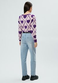 Lila Herzmuster auf einem flauschigen, hellrosa Pullover mit gerippten Bündchen. Hellblaue High-Waist-Jeans und schwarze klobige Plateausneaker.